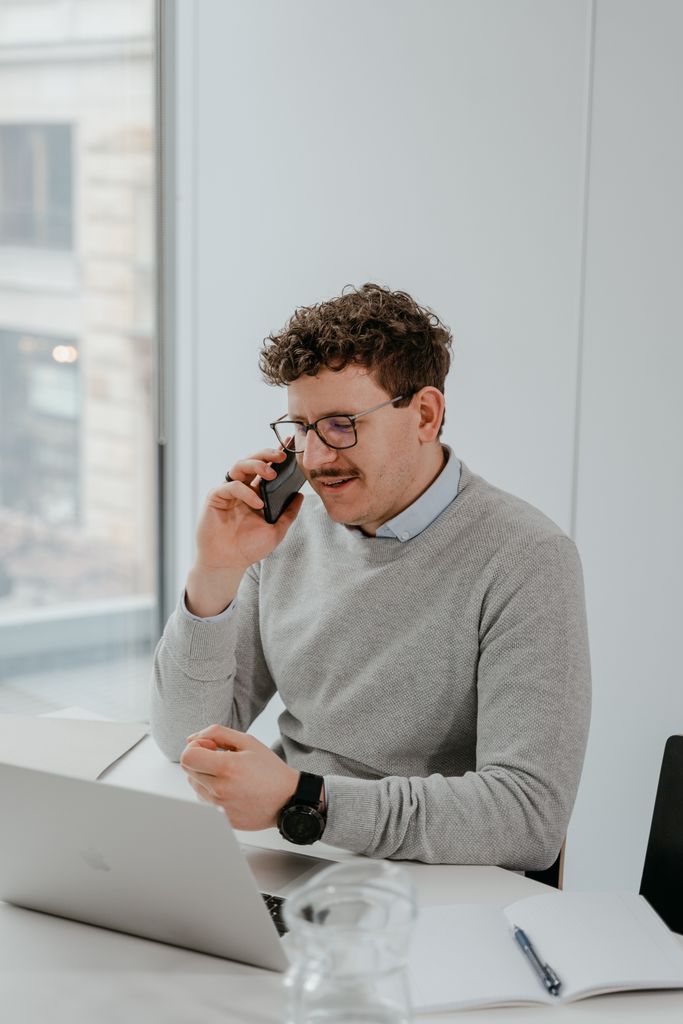 Young adult man in a gray sweater on a phone call, working on a laptop in a modern office.